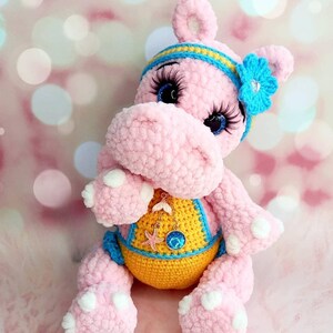 May include: A pink crocheted hippo plush toy with blue eyes, long eyelashes, and a blue and yellow headband. The hippo wears a yellow and blue swimsuit and a necklace with a starfish charm. The plush toy has white spots on its feet.
