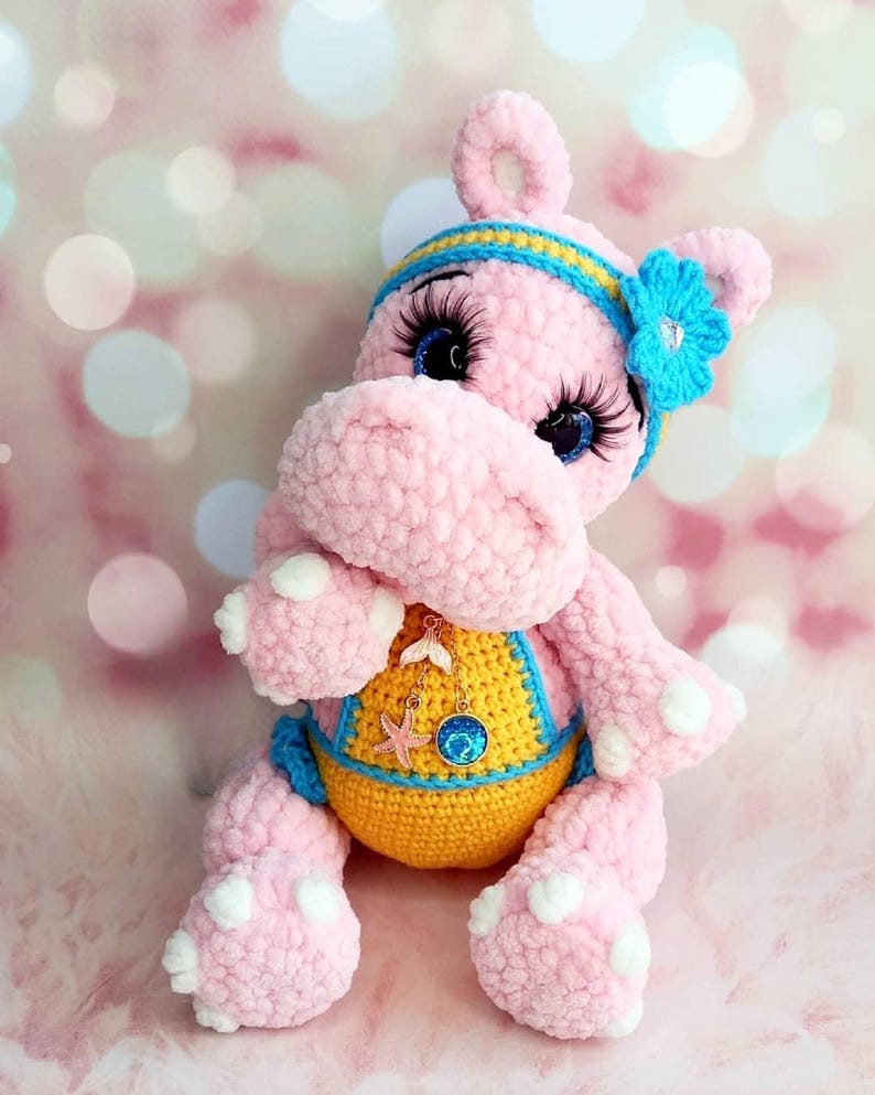 May include: A pink crocheted hippo plush toy with blue eyes, long eyelashes, and a blue and yellow headband. The hippo wears a yellow and blue swimsuit and a necklace with a starfish charm. The plush toy has white spots on its feet.