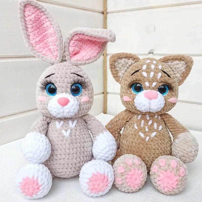 May include: Two handmade crochet stuffed animals: a bunny and a cat. The bunny is light gray with pink inner ears and paws. The cat is brown with white markings. Both have blue eyes and long eyelashes. The bunny is approximately 10 inches tall.