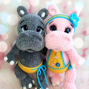 Op de afbeelding: Twee gehaakte nijlpaardspeeltjes. Eén is grijs met gele en blauwe details, de andere is roze met een blauw en gele hoofdband en een decoratieve slabbetje. Beide hebben blauwe ogen en witte accenten op hun voeten.