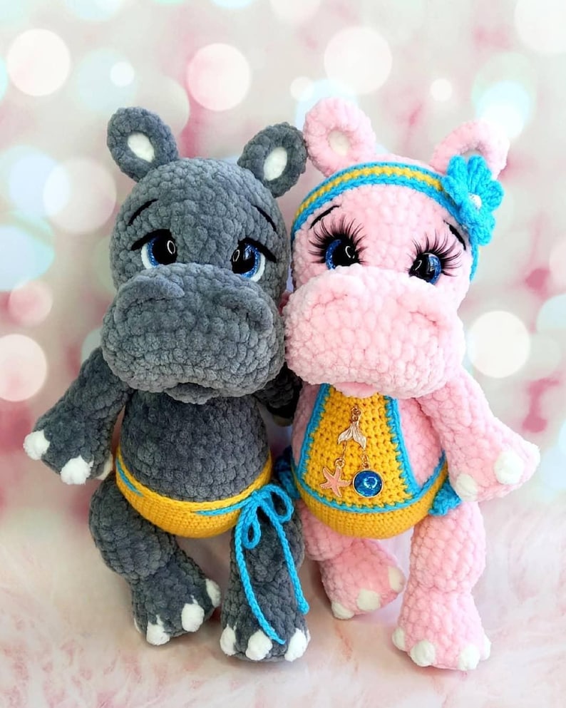 May include: Two plush hippopotamus toys. One is gray with a yellow and blue accent, the other is pink with a blue and yellow headband and a decorative bib. Both have blue eyes and white accents on their feet.