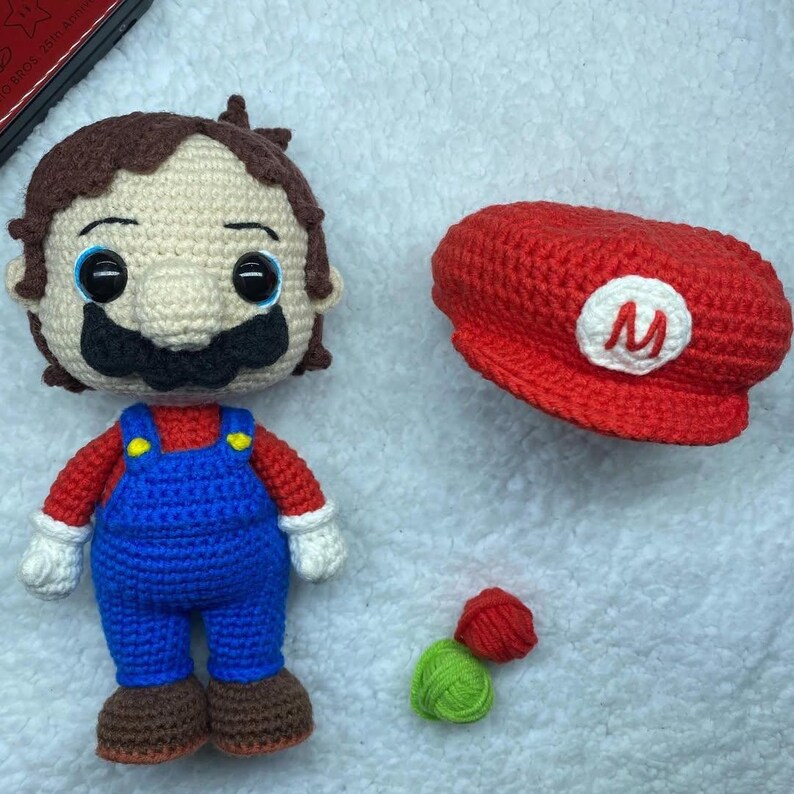 May include: A handmade crochet Mario doll with a red shirt, blue overalls, and a brown mustache. A red hat with a white "M" emblem sits next to the doll. Additional small crochet pieces in red and green are also present.