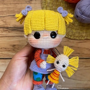 Puede incluir: Muñeca de ganchillo hecha a mano con cabello amarillo, lazos morados y ojos negros grandes. La muñeca viste un vestido morado, mangas naranjas y pantalones azules con lunares verdes. Se sostiene una muñeca más pequeña. El nombre "Sienna" está escrito abajo a la derecha.