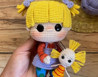 May include: A handmade crochet doll with yellow hair, purple bows, and large black eyes. The doll wears a purple dress, orange sleeves, and blue pants with green polka dots. A smaller doll is held in its hand. The name "Sienna" is written on the bottom right.