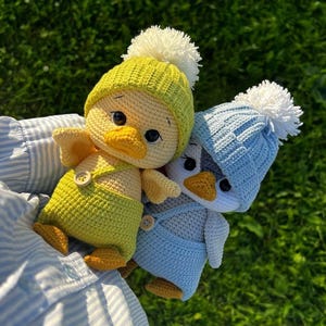 May include: Two crocheted duck toys, one yellow and one blue, each wearing a matching hat with a white pom-pom. The yellow duck has yellow dungarees. The ducks are set against a green background.