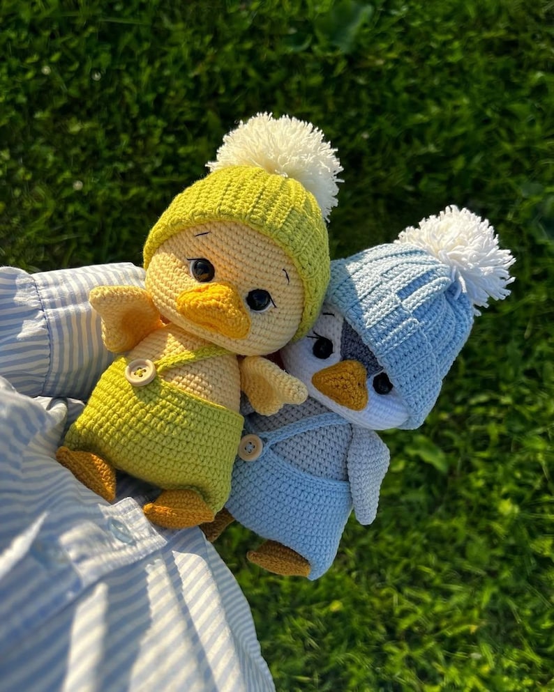 May include: Two crocheted duck toys, one yellow and one blue, each wearing a matching hat with a white pom-pom. The yellow duck has yellow dungarees. The ducks are set against a green background.