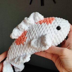 May include: A handmade, plush koi fish toy. The fish is primarily white with orange markings and a black eye. The soft, textured material gives the fish a cuddly appearance. The toy is held in a person's hands.