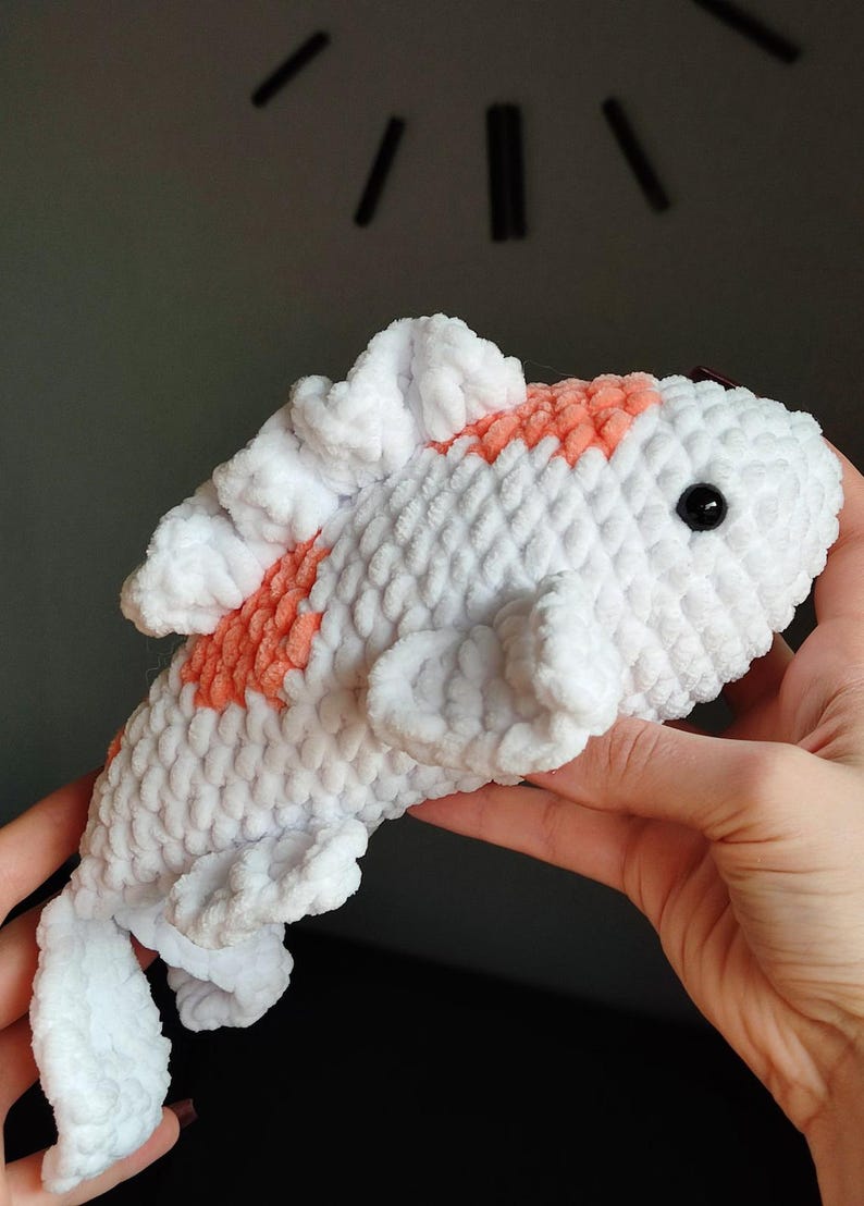 May include: A handmade, plush koi fish toy. The fish is primarily white with orange markings and a black eye. The soft, textured material gives the fish a cuddly appearance. The toy is held in a person's hands.