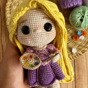 May include: A handmade crochet doll with yellow hair and a purple dress, holding a paintbrush and a colourful palette. The doll has large black eyes and rosy cheeks. The doll is approximately 15 cm tall.