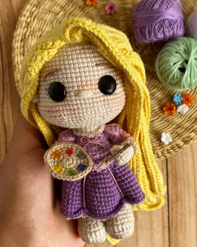 May include: A handmade crochet doll with yellow hair and a purple dress, holding a paintbrush and a colorful palette. The doll has large black eyes and rosy cheeks. The doll is approximately 6 inches tall.