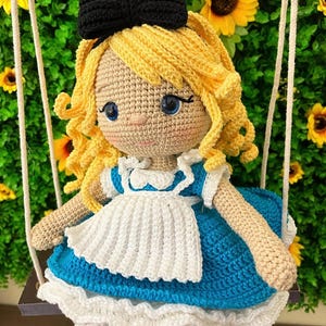 Puede incluir: Mu&ntilde;eca de ganchillo de una ni&ntilde;a con cabello rubio rizado, ojos azules y un lazo negro. Lleva un vestido azul y blanco y est&aacute; sentada en un columpio. El fondo presenta girasoles.