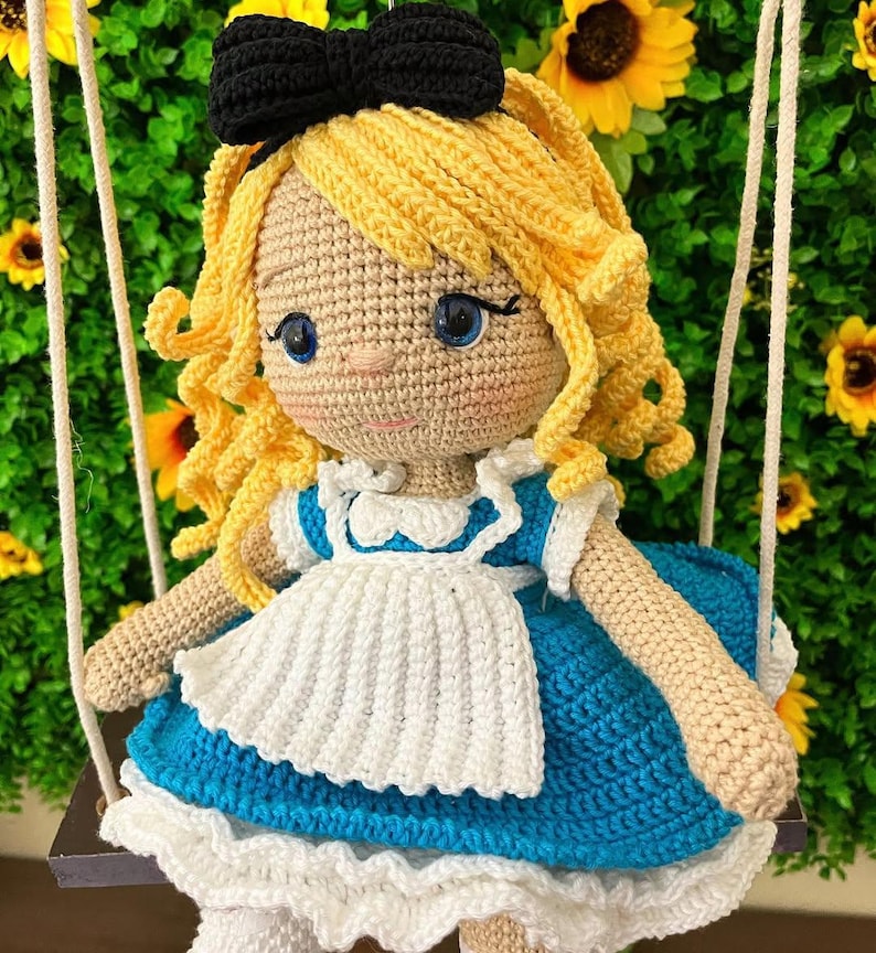 Puede incluir: Mu&ntilde;eca de ganchillo de una ni&ntilde;a con cabello rubio rizado, ojos azules y un lazo negro. Lleva un vestido azul y blanco y est&aacute; sentada en un columpio. El fondo presenta girasoles.