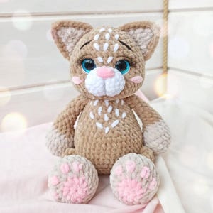 May include: A handmade crochet cat doll in shades of brown, white, and pink. The cat has large blue eyes, a pink nose, and white markings. The paws are pink with gray pads. The doll is sitting on a pink surface.