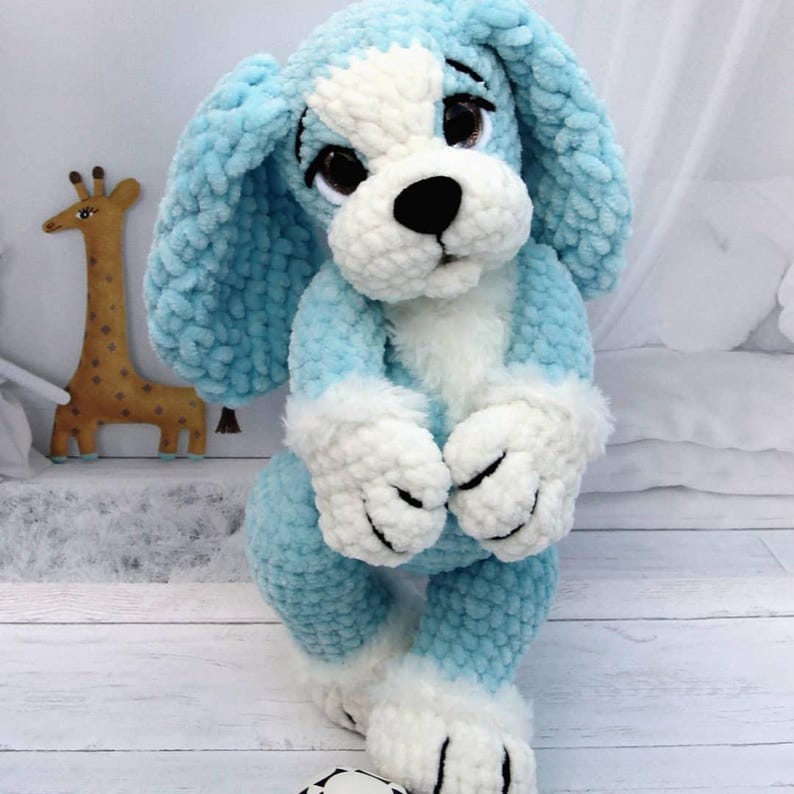 May include: A handmade plush dog toy in light blue and white. The dog has large floppy ears, a white face, and white paws. The toy is made of soft, textured yarn and has black eyes and a black nose. A giraffe toy is in the background.