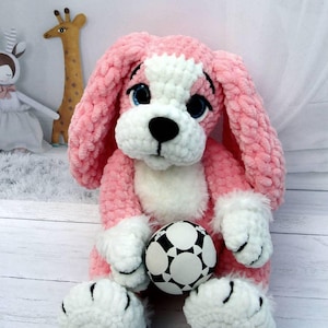 May include: A handmade pink and white plush dog with blue eyes, a black nose, and a black and white soccer ball. The dog has long, floppy ears and white paws. The plush toy is sitting on a white surface.