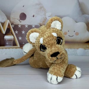 Puede incluir: Un peluche de cachorro de le&oacute;n en tonos marr&oacute;n y blanco. El le&oacute;n tiene un cuerpo texturizado y difuso, con detalles blancos en las orejas y las patas. Tiene ojos grandes y expresivos con brillo dorado y una nariz negra. El juguete est&aacute; tumbado sobre una superficie clara.