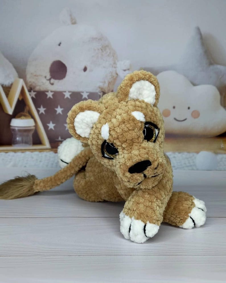 Puede incluir: Un peluche de cachorro de le&oacute;n en tonos marr&oacute;n y blanco. El le&oacute;n tiene un cuerpo texturizado y difuso, con detalles blancos en las orejas y las patas. Tiene ojos grandes y expresivos con brillo dorado y una nariz negra. El juguete est&aacute; tumbado sobre una superficie clara.
