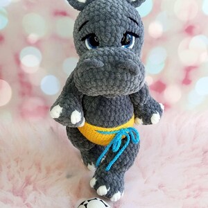 May include: A handmade gray crochet hippo toy with blue eyes, white accents, and a yellow waistband tied with blue yarn. A black and white soccer ball sits at the hippo's feet. The hippo is a plush toy.