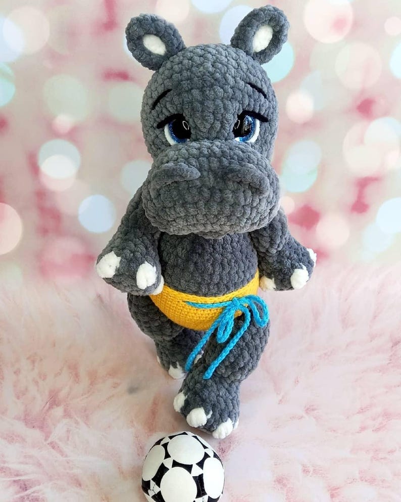 May include: A handmade gray crochet hippo toy with blue eyes, white accents, and a yellow waistband tied with blue yarn. A black and white soccer ball sits at the hippo's feet. The hippo is a plush toy.