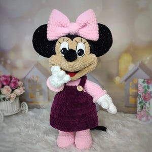 Puede incluir: Mu&ntilde;eca de peluche de Minnie Mouse hecha a mano. La mu&ntilde;eca tiene orejas negras, cara color canela y un lazo rosa. Lleva un su&eacute;ter rosa, un vestido morado oscuro, guantes blancos y zapatos rosas. La mu&ntilde;eca est&aacute; sobre una superficie blanca.