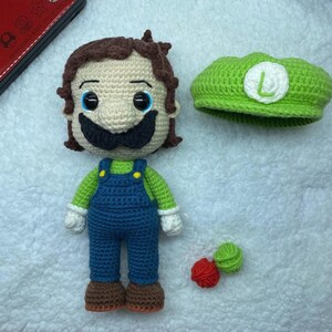May include: A handmade crochet doll of a character with a large black mustache, green shirt, and blue overalls. A green hat with a white "L" is next to the doll. Two small balls of yarn are also present.