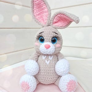 May include: A handmade crochet bunny plush toy. The bunny is light gray with pink accents on the inner ears, nose, and feet. It has large blue eyes with long eyelashes and white fluffy paws. The bunny is sitting on a pink surface.