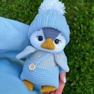 May include: A crocheted penguin toy. The penguin has a white face, grey head, and yellow beak and feet. It is wearing a light blue hat with a white pom-pom and light blue dungarees with a wooden button.