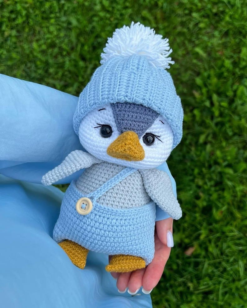 May include: A crocheted penguin toy. The penguin has a white face, grey head, and yellow beak and feet. It is wearing a light blue hat with a white pom-pom and light blue dungarees with a wooden button.