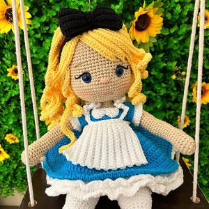 Puede incluir: Mu&ntilde;eca de ganchillo hecha a mano de una ni&ntilde;a con cabello rubio rizado y un lazo negro. La mu&ntilde;eca lleva un vestido azul y blanco y est&aacute; sentada en un columpio. El fondo presenta girasoles y vegetaci&oacute;n.