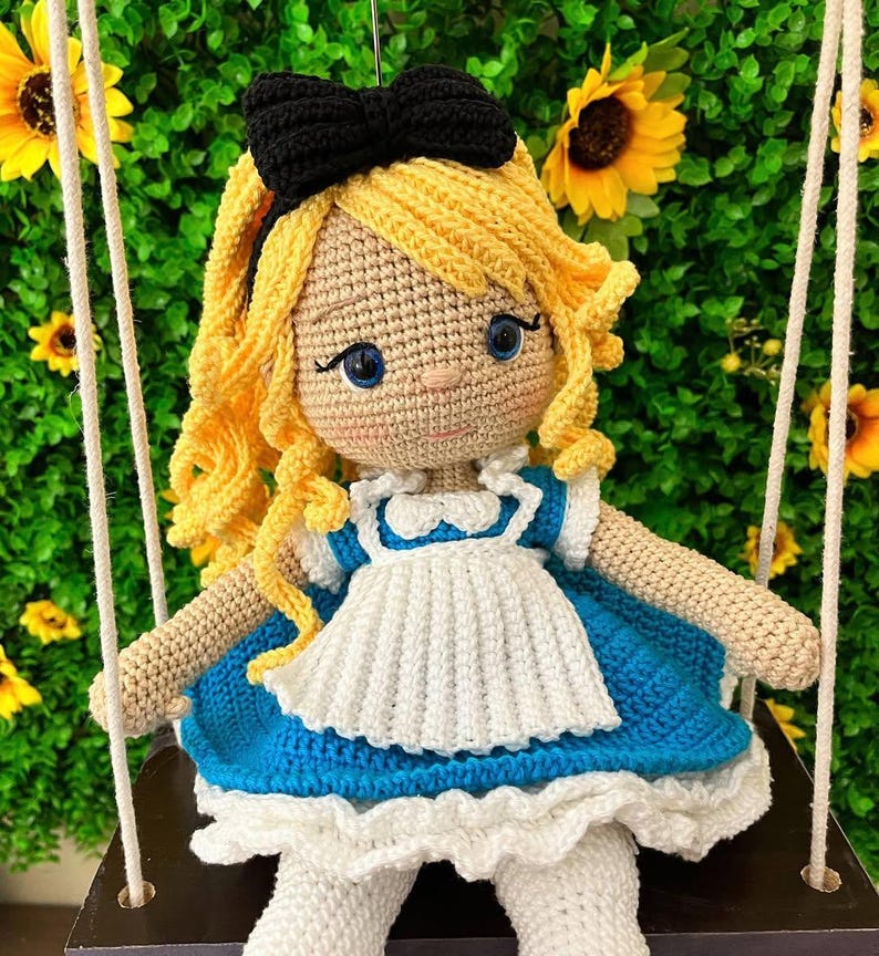 Puede incluir: Mu&ntilde;eca de ganchillo hecha a mano de una ni&ntilde;a con cabello rubio rizado y un lazo negro. La mu&ntilde;eca lleva un vestido azul y blanco y est&aacute; sentada en un columpio. El fondo presenta girasoles y vegetaci&oacute;n.