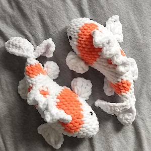 May include: Two handmade crochet koi carp in white and orange. The fish have black eyes and are lying on a grey surface. The koi carp are a soft, plush design.