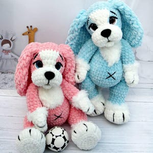 May include: Two plush crochet dog toys. One is pink with white accents and a black nose, the other is blue with white accents and a black nose. Both have floppy ears and embroidered details. A soccer ball is next to the pink dog.