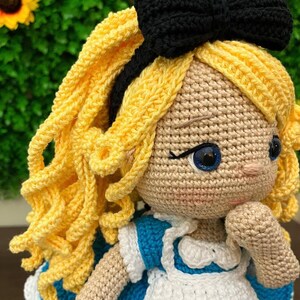 Puede incluir: Mu&ntilde;eca de ganchillo hecha a mano con cabello rizado amarillo, ojos azules y un lazo negro. La mu&ntilde;eca lleva un vestido azul y blanco. La piel de la mu&ntilde;eca es de color canela claro. La mu&ntilde;eca es una representaci&oacute;n de un personaje de cuento infantil.