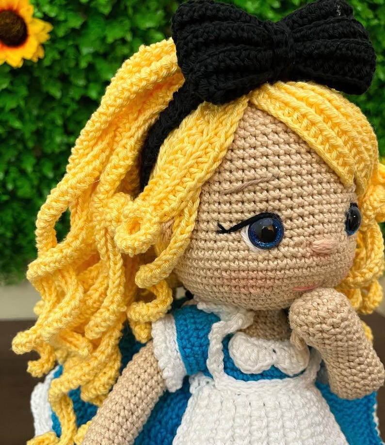 Puede incluir: Mu&ntilde;eca de ganchillo hecha a mano con cabello rizado amarillo, ojos azules y un lazo negro. La mu&ntilde;eca lleva un vestido azul y blanco. La piel de la mu&ntilde;eca es de color canela claro. La mu&ntilde;eca es una representaci&oacute;n de un personaje de cuento infantil.