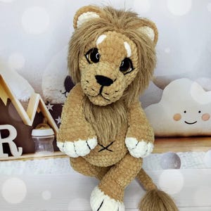 Puede incluir: Un juguete de le&oacute;n de peluche con un cuerpo marr&oacute;n, una melena marr&oacute;n y detalles blancos en las patas. El le&oacute;n tiene ojos grandes y expresivos y una nariz y boca negras cosidas. El juguete est&aacute; sentado con las piernas cruzadas y su cola es visible.