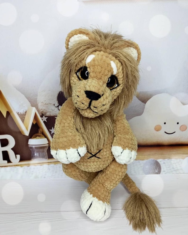 Puede incluir: Un juguete de le&oacute;n de peluche con un cuerpo marr&oacute;n, una melena marr&oacute;n y detalles blancos en las patas. El le&oacute;n tiene ojos grandes y expresivos y una nariz y boca negras cosidas. El juguete est&aacute; sentado con las piernas cruzadas y su cola es visible.