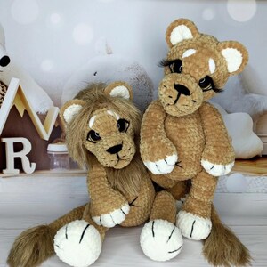 Puede incluir: Dos peluches de le&oacute;n hechos a mano con ganchillo. Los leones son marrones con detalles blancos en las patas y las orejas. Un le&oacute;n tiene melena y el otro tiene largas pesta&ntilde;as. Los juguetes est&aacute;n sobre una superficie blanca.