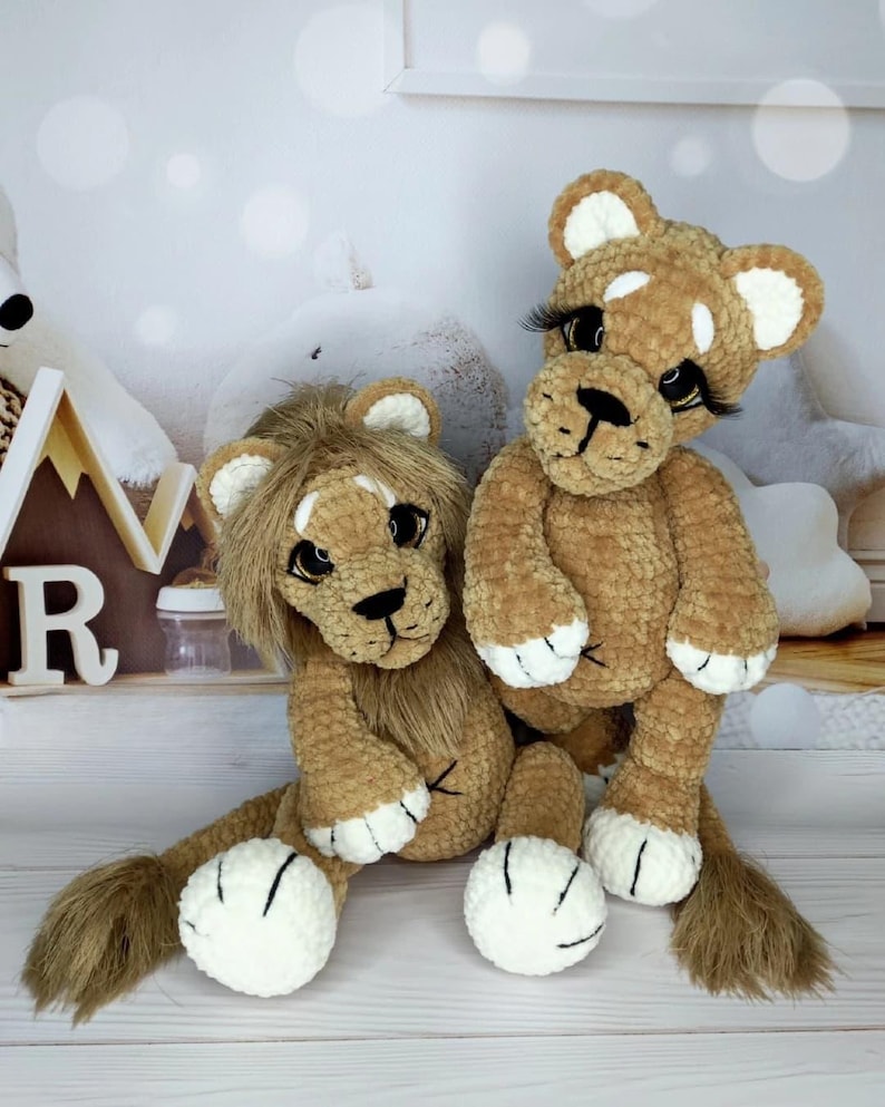 Puede incluir: Dos peluches de le&oacute;n hechos a mano con ganchillo. Los leones son marrones con detalles blancos en las patas y las orejas. Un le&oacute;n tiene melena y el otro tiene largas pesta&ntilde;as. Los juguetes est&aacute;n sobre una superficie blanca.