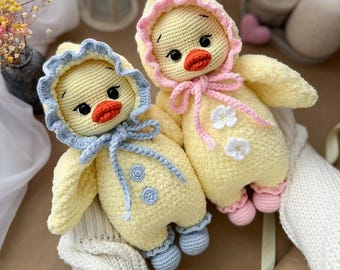 May include: Two handmade crochet duckling dolls. The dolls are yellow with blue and pink accents. Each duckling wears a hooded jumpsuit with a matching bonnet. The ducklings have black eyes and orange beaks. One has a blue bow, the other a pink bow.