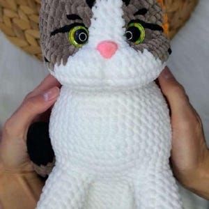 以下が含まれることがあります： 手作りのかぎ針編みの猫の人形。白い体と、グレー、茶色、オレンジ色のアクセントが施された頭部が特徴です。猫は緑色の目、ピンクの鼻、ピンクの肉球を持っています。人形は手で持たれています。