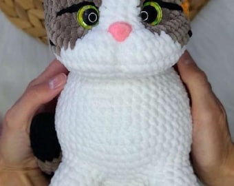 May include: A handmade crochet cat doll with a white body and a head featuring gray, brown, and orange accents. The cat has green eyes, a pink nose, and pink paw pads. The doll is held by hands.