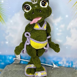 May include: A handmade green plush frog wearing a yellow and purple bikini, sandals, and a silver crown. The frog has large, expressive eyes, a pink tongue, and is posed with arms out. The background shows a beach scene.