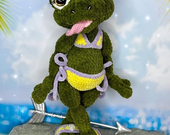 May include: A handmade green plush frog wearing a yellow and purple bikini, sandals, and a silver crown. The frog has large, expressive eyes, a pink tongue, and is posed with arms out. The background shows a beach scene.