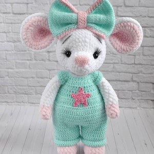 Może przedstawiać: Ręcznie robiona szydełkowa lalka myszy z białym korpusem, różowymi i białymi uszami oraz miętową kokardą. Mysz nosi miętowy kombinezon z różową gwiazdą. Lalka to miękka zabawka.