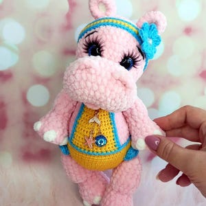 May include: A handmade pink crochet hippo doll with a yellow and blue swimsuit. The hippo has a blue and yellow headband with a blue flower, and long eyelashes. A small charm hangs from the hippo's neck.