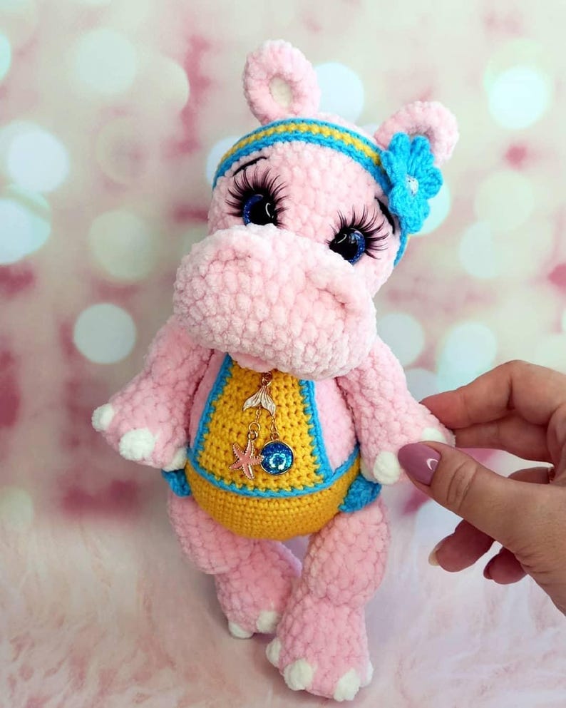 May include: A handmade pink crochet hippo doll with a yellow and blue swimsuit. The hippo has a blue and yellow headband with a blue flower, and long eyelashes. A small charm hangs from the hippo's neck.