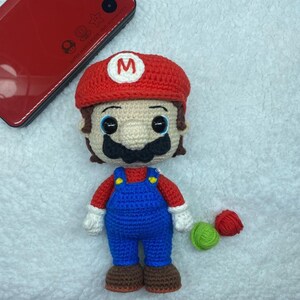 May include: A handmade crochet doll of the character Mario, wearing a red hat with a white "M", a red shirt, blue overalls, and brown shoes. The doll has a black mustache and brown hair. A red game console is in the background.