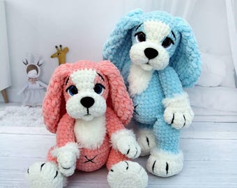 May include: Two plush crochet dog toys. One is pink with white accents, the other is blue with white accents. Both have black eyes and noses. The dogs are sitting on a light wood surface.