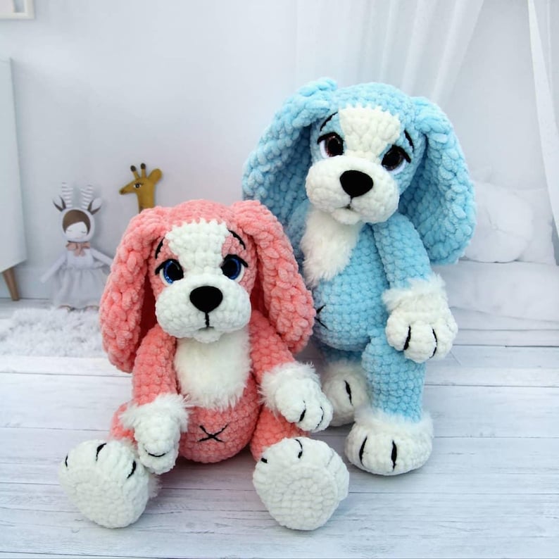 May include: Two plush crochet dog toys. One is pink with white accents, the other is blue with white accents. Both have black eyes and noses. The dogs are sitting on a light wood surface.
