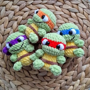 May include: Four handmade crochet turtle toys in green, yellow, and brown. Each turtle has a different coloured headband: orange, blue, red, and purple. The turtles have black eyes and are arranged on a woven, light brown surface.